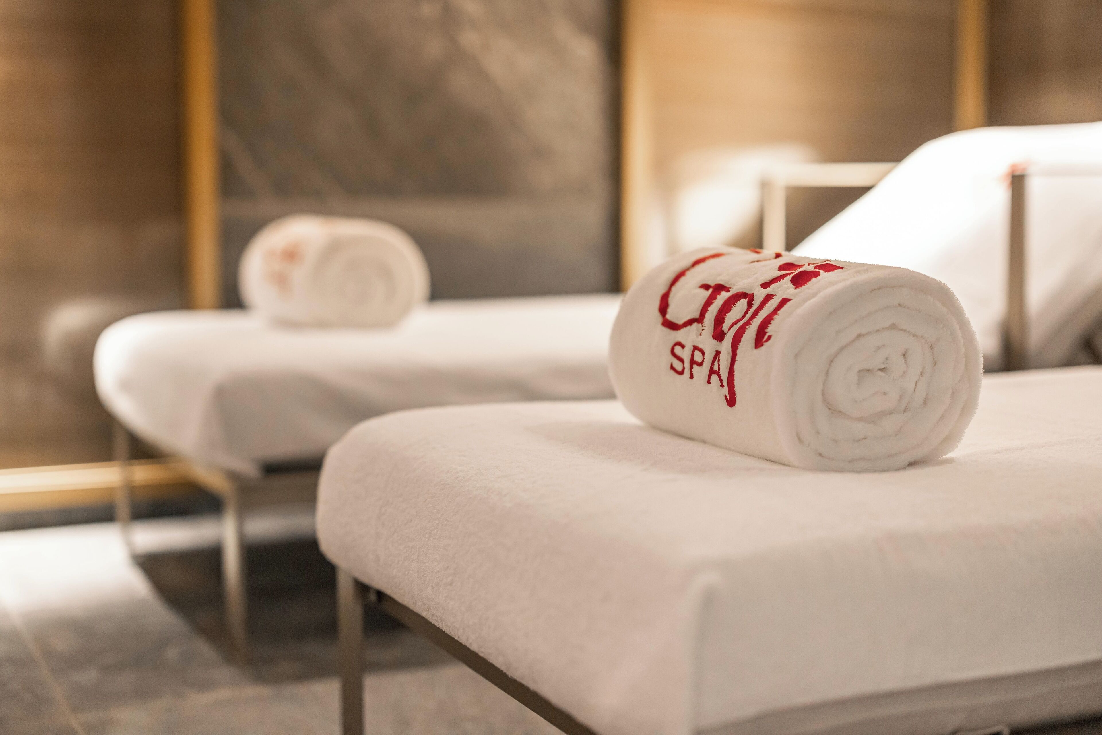 Two spa beds with towels rolled up, one with "Spa" embroidered on it, in a modern room with a marble floor and a large mirror.