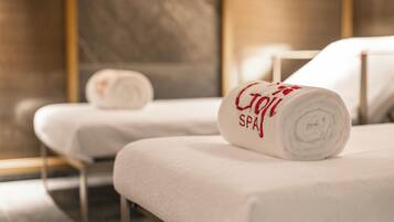 Two spa beds with towels rolled up, one with "Spa" embroidered on it, in a modern room with a marble floor and a large mirror.