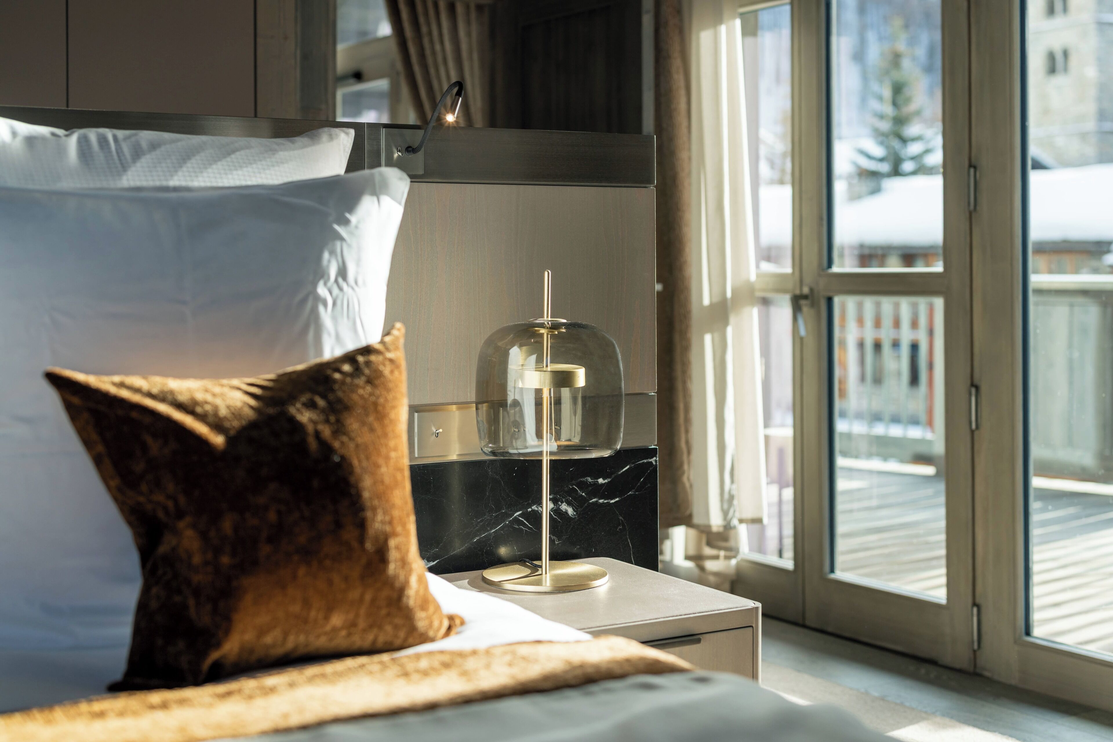 Habitación de hotel con cama, mesita de noche, lámpara con cupula de vidrio y vistas a un paisaje nevado.