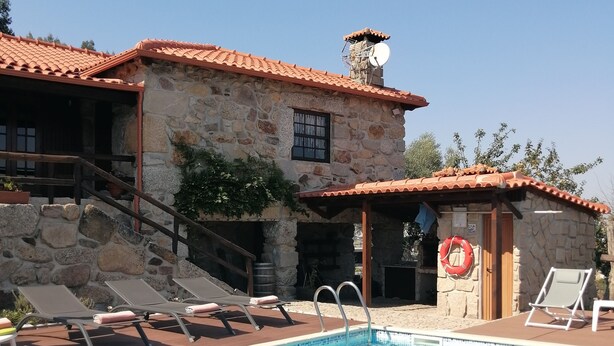 Ein Steinhaus mit Ziegeldach, einem Schwimmbecken mit Liegestühlen und Schwimmreifen sowie einem Rettungsring an der Wand.