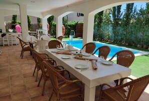 Área de refeições ao ar livre com vista para a piscina, cadeiras de vime e uma mesa posta para quatro pessoas.