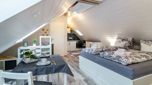 A compact, well-lit attic bedroom with a dining area, kitchenette, and a cozy bed with patterned bedding.