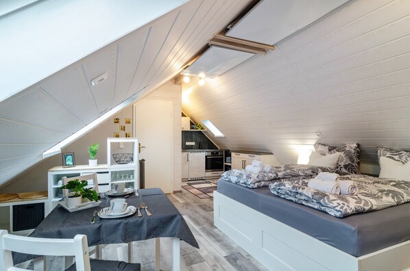 A compact, well-lit attic bedroom with a dining area, kitchenette, and a cozy bed with patterned bedding.