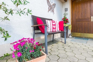 A bench with red cushions, a potted plant with pink flowers, and a wooden door.