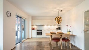 Une cuisine moderne avec des meubles blancs, une table à manger avec des chaises, un luminaire suspendu et une horloge au mur.
