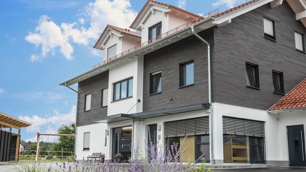 Un immeuble résidentiel moderne à deux étages, doté d’un balcon, de grandes fenêtres et d’un espace extérieur aménagé et couvert.