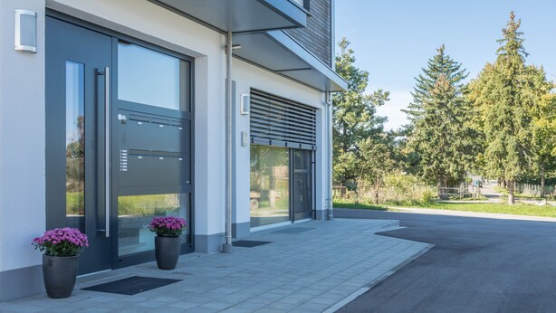 A modern building with large glass doors, a metal entrance, and potted plants on the sidewalk.