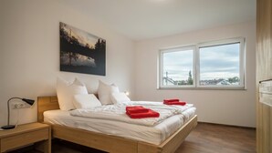 A bedroom with a wooden bed, white bedding, a nightstand, a desk, a window with a view, and a painting on the wall.