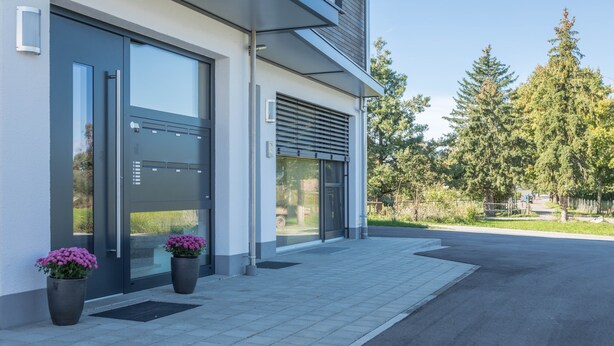 Un edificio moderno con grandes puertas de vidrio, plantas en macetas y un sendero pavimentado.