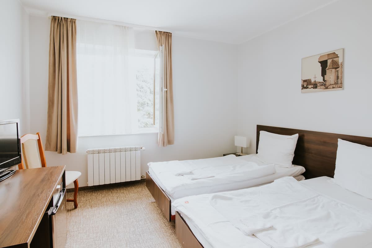Habitación de hotel con dos camas, un escritorio de madera, una silla, un radiador y una ventana con cortinas.