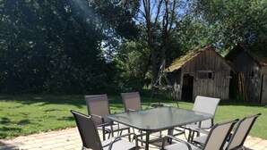 Une terrasse avec une table et des chaises en verre, un sol en briques et une structure en bois en arrière-plan.