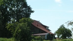 Une maison de campagne avec un toit rouge, entourée d’un champ verdoyant et d’arbres.