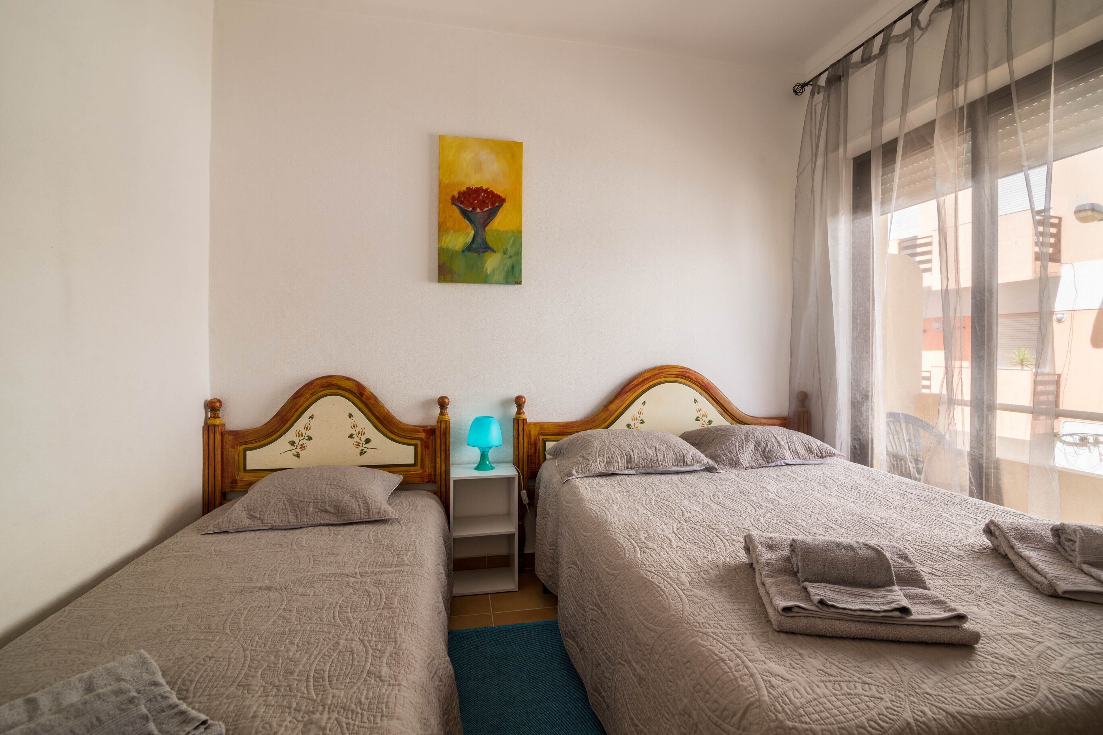 A hotel room with two beds, a nightstand with a lamp, a painting on the wall, and a view of the outside.