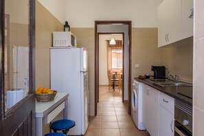 A narrow kitchen with a refrigerator, microwave, sink, and a dining area in the background.