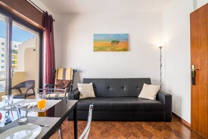 Una sala de estar moderna con un sofá negro, una mesa de comedor con sillas, un cuadro en la pared y vistas a través de una ventana.