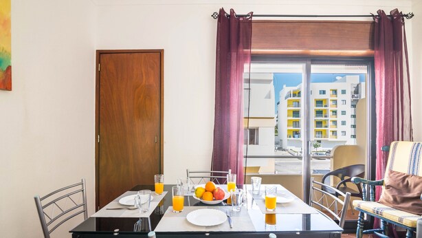 A dining area with a table set for two, a window with curtains, and a view of buildings outside. - "Beach Residence 21" with Balcony, Barbecue, Wi-Fi & Cable TV; Parking Available (Armação de Pera)
