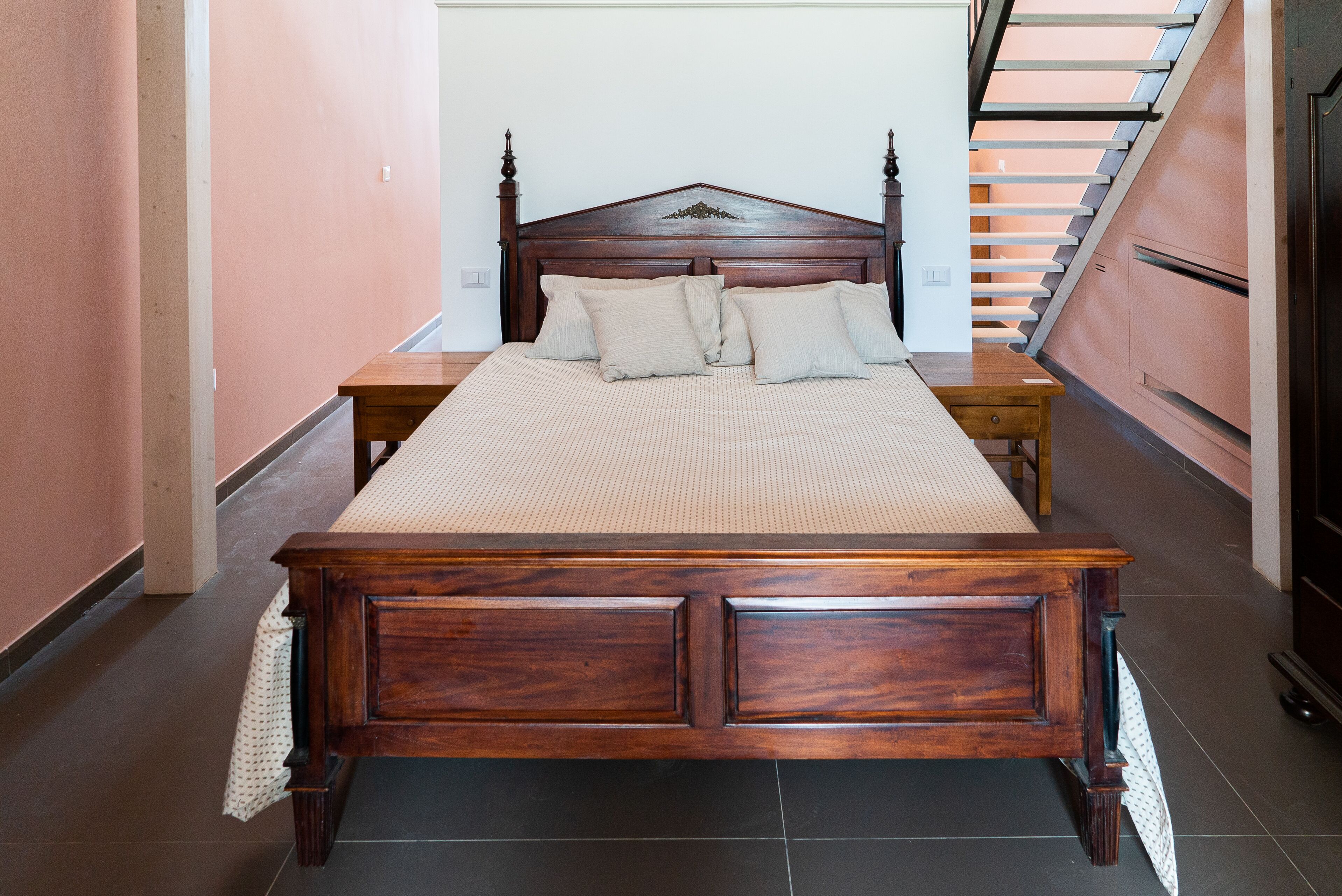A bedroom with a large wooden bed, a nightstand, and a staircase in the background.