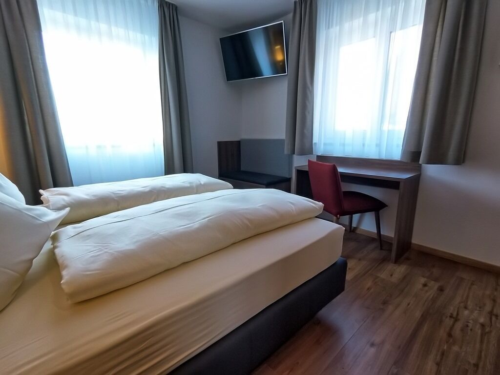 A hotel room with a bed, a desk, a chair, and a TV mounted on the wall.