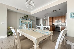 A dining area with a long table, white chairs, and a blue table runner. There is a kitchen area with modern appliances and wooden cabinets.