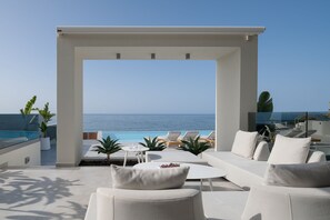 A modern outdoor lounge area with a view of the ocean, featuring white furniture, a glass balustrade, and potted plants. - Hermes Grand Luxury Beachfront Villa & Spa-Private Beach, Turkish Hammam & Sauna (Skaleta, Rethymno)