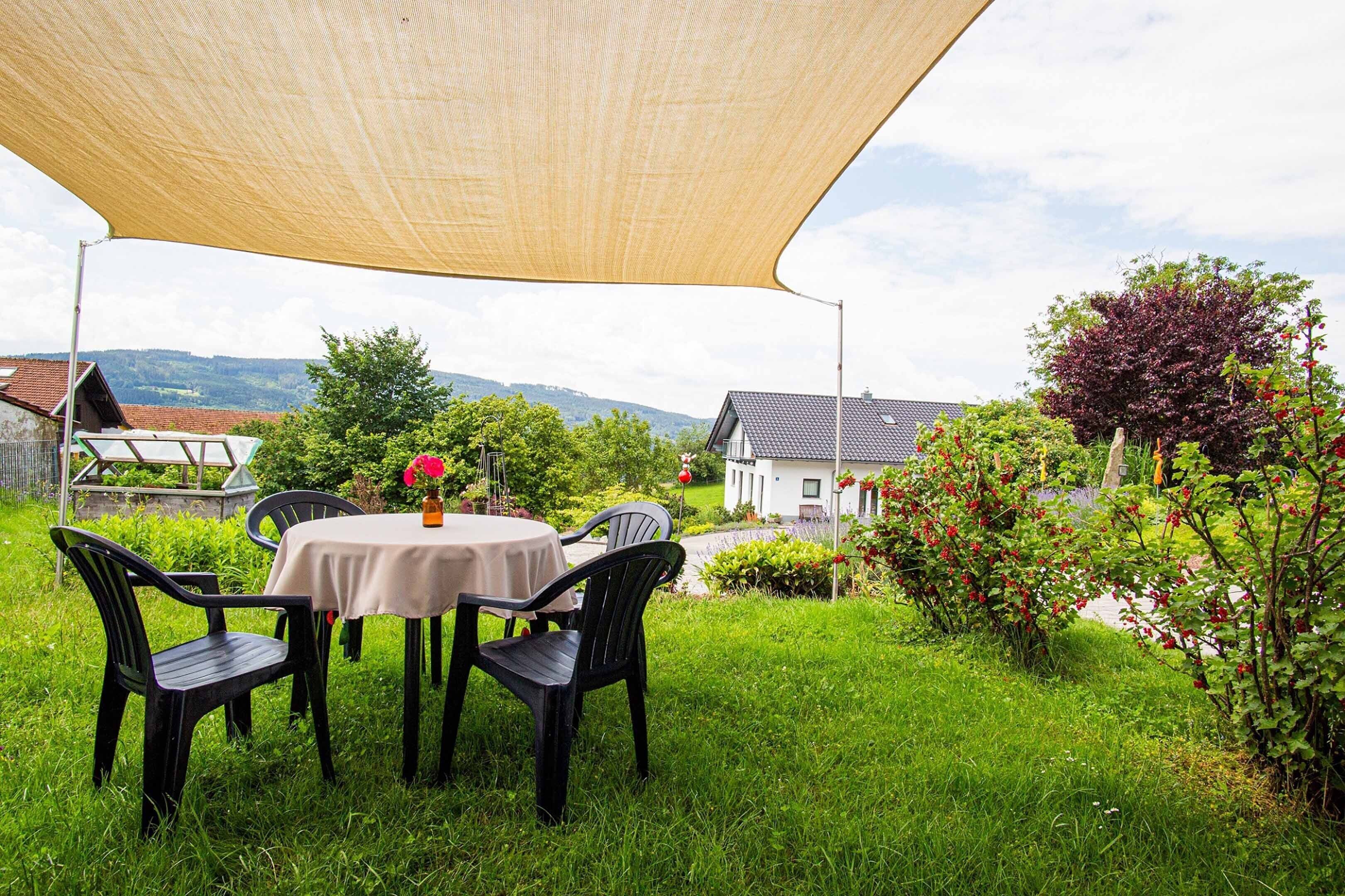 An outdoor dining area with a table and chairs under a shade sail, surrounded by lush greenery and a view of houses in the background.