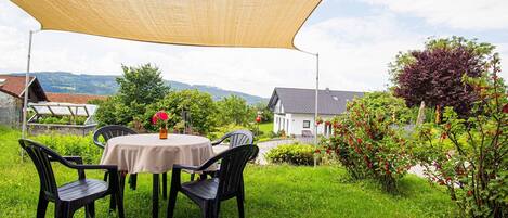 An outdoor dining area with a table and chairs under a shade sail, surrounded by lush greenery and a view of houses in the background.