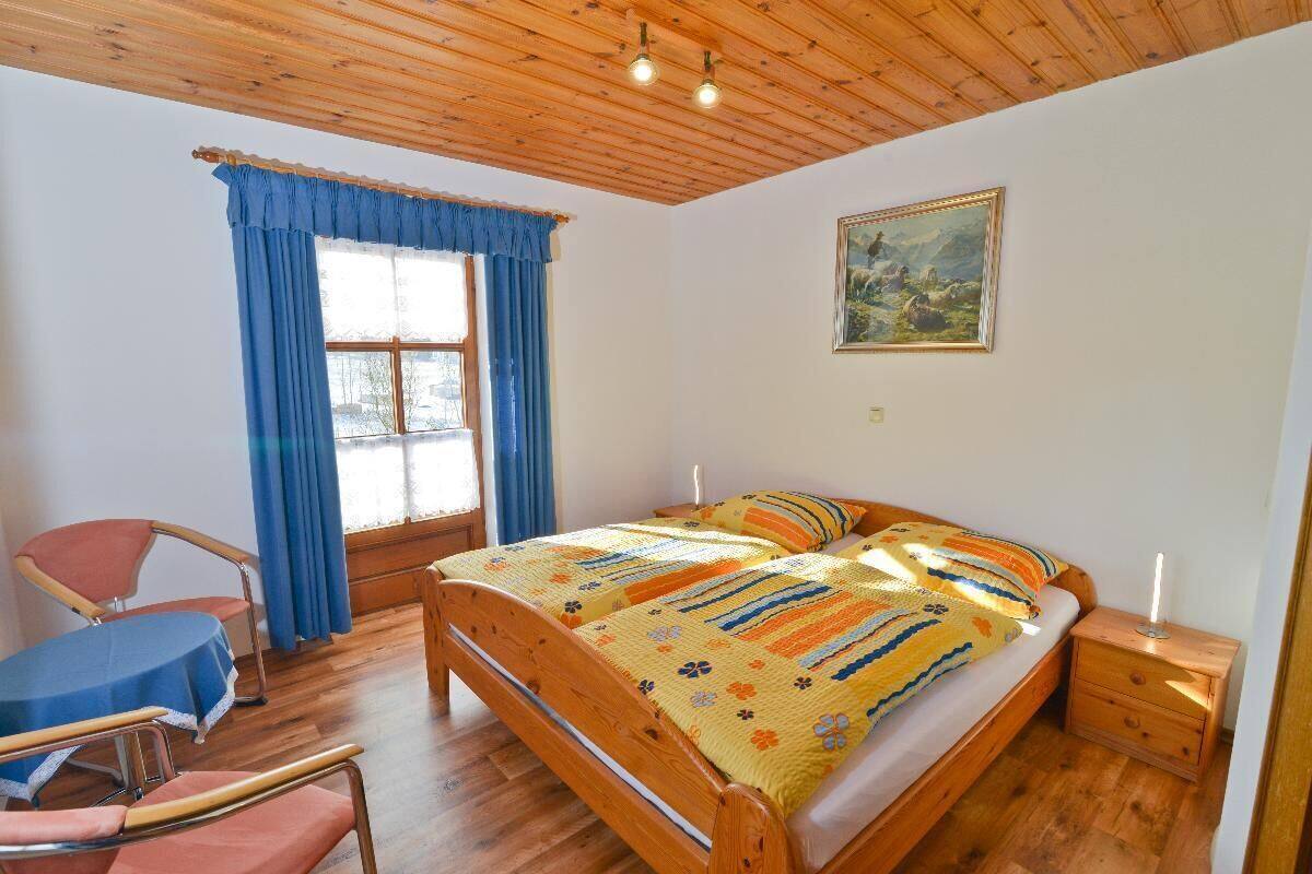 Ein Schlafzimmer mit einem hölzernen Bett, einem blauen Sessel, einem kleinen Tisch und einem Fenster mit blauen Vorhängen.