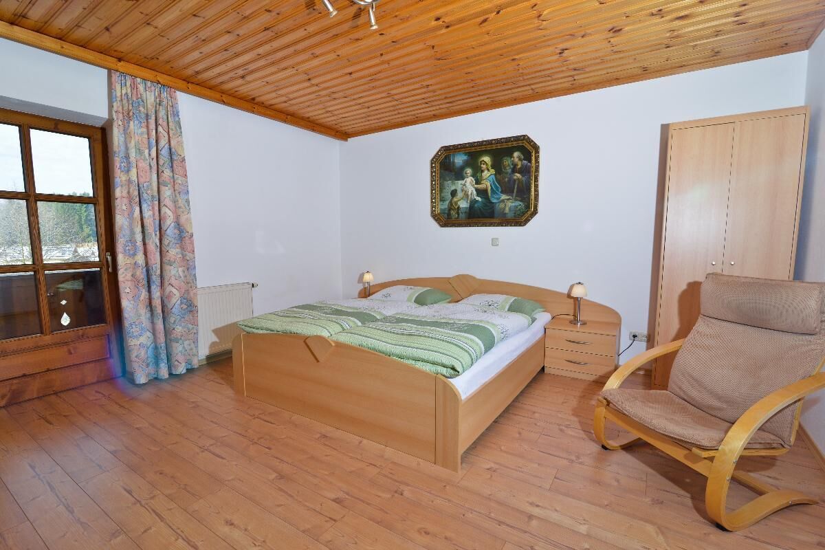 Un dormitorio con una cama de madera, una silla, un aparador, una ventana con cortinas y un cuadro en la pared.