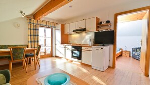 Een kleine keuken met witte kastjes, een fornuis en een vaatwasser. Er is een eethoek met een tafel en stoelen, en een zithoek met een bank en een kom op de vloer.
