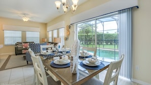 A dining area with a glass table, white chairs, and a chandelier. There is a sofa and a window with blinds in the background. - South-Facing Private Pool • Gated Resort • Resort Amenities • Near Disney (Kissimmee)