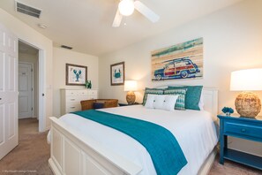 A bedroom with a large bed, a ceiling fan, a nightstand, a lamp, and artwork on the wall.