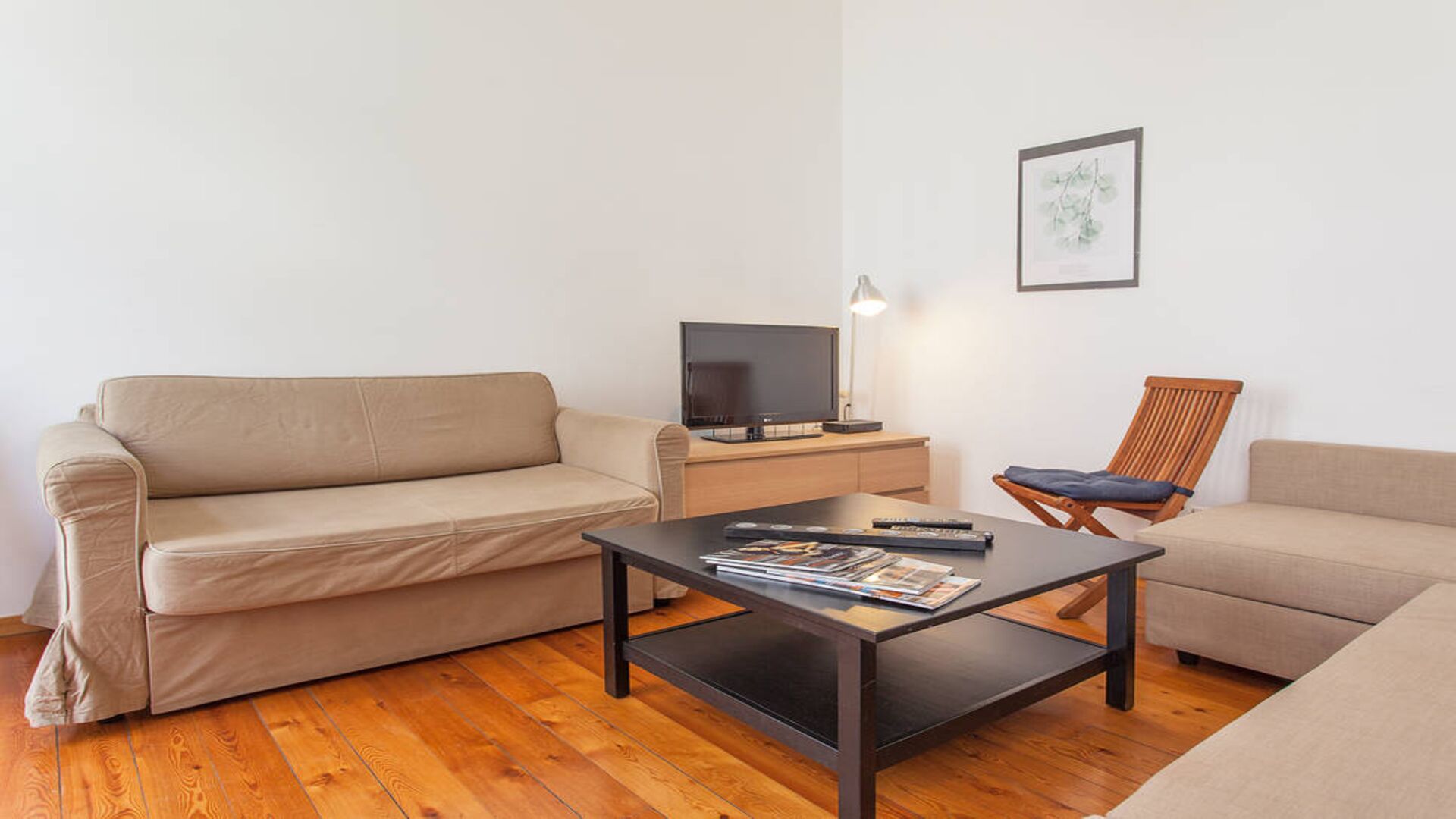 A living room with a beige sofa, a wooden coffee table, a TV on a wooden stand, and a framed picture on the wall.