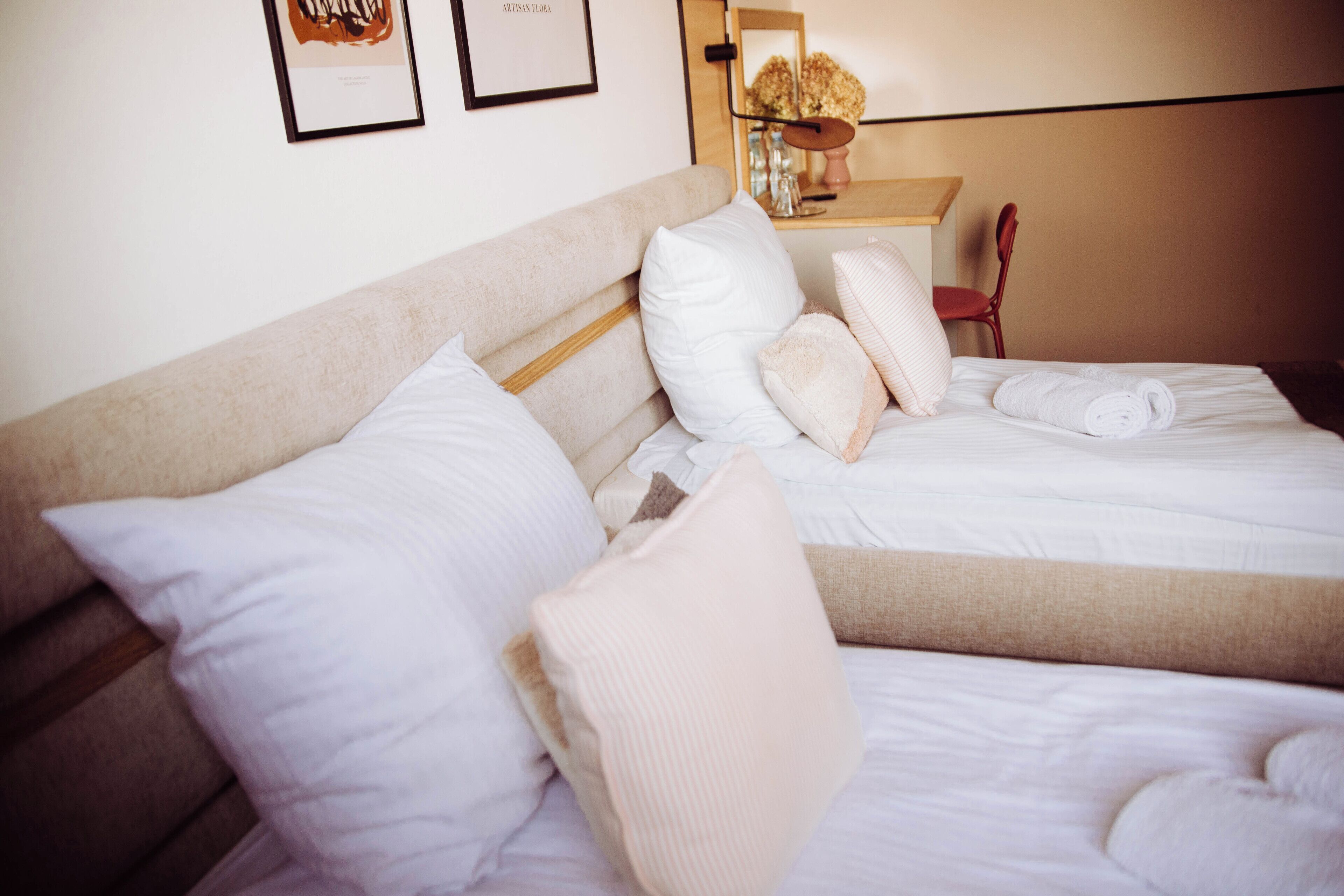 A neatly made bed with white and beige bedding, a wooden headboard, and a mirror above it reflecting a vase with flowers.