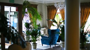 Un hall d’entrée avec un fauteuil bleu, des plantes en pot, un lustre et un panneau indiquant la sortie.