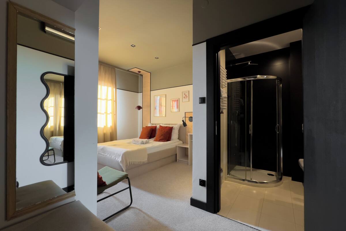 Una habitación de hotel moderna con cama, mesita de noche, espejo, cabina de ducha y vista al pasillo.