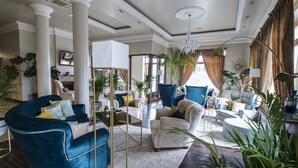 Un salon spacieux avec un grand fauteuil bleu, un fauteuil blanc et un canapé bleu. On y trouve également plusieurs plantes en pot et un lustre.