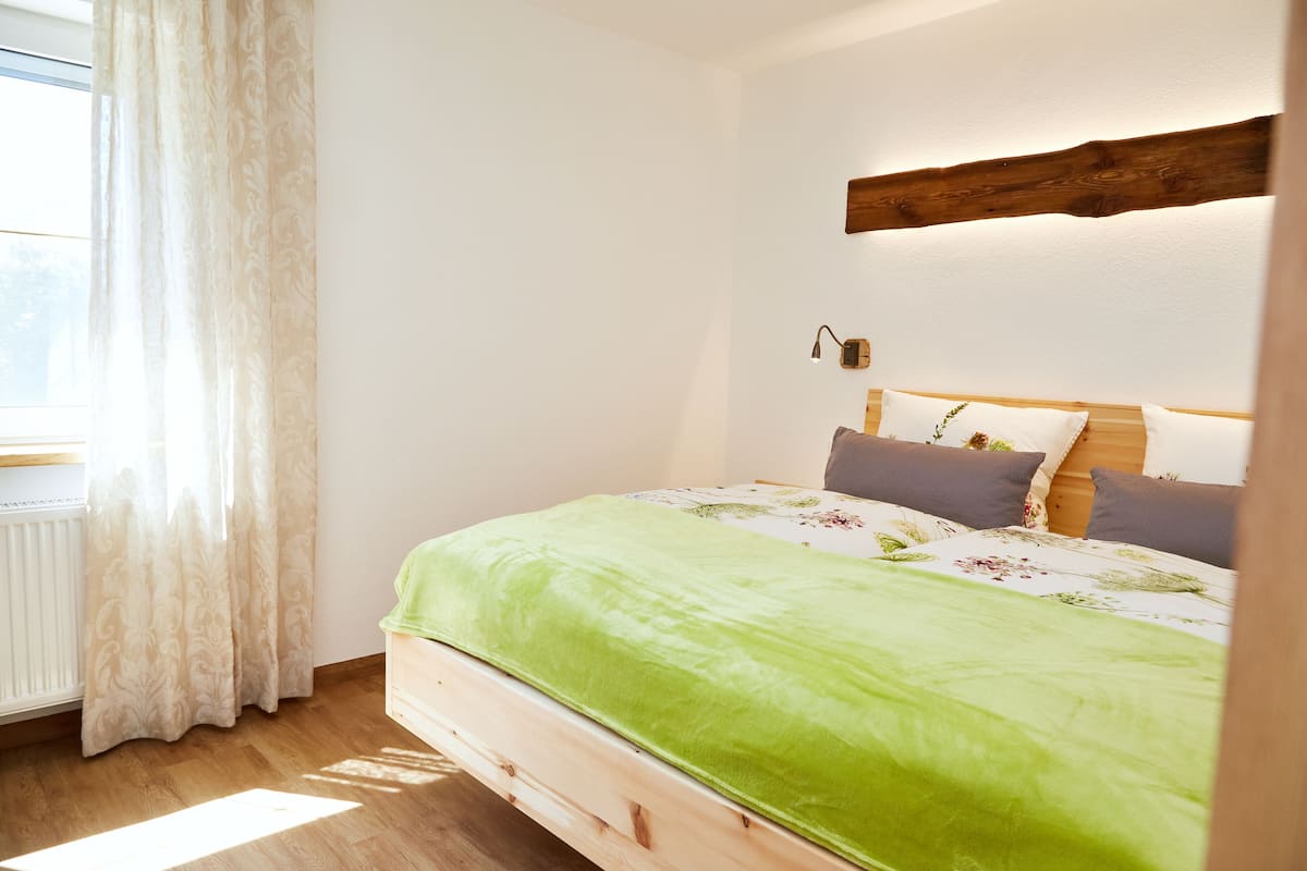 Un dormitorio con estructura de cama de madera, colcha verde y ventana con cortinas de gasa.