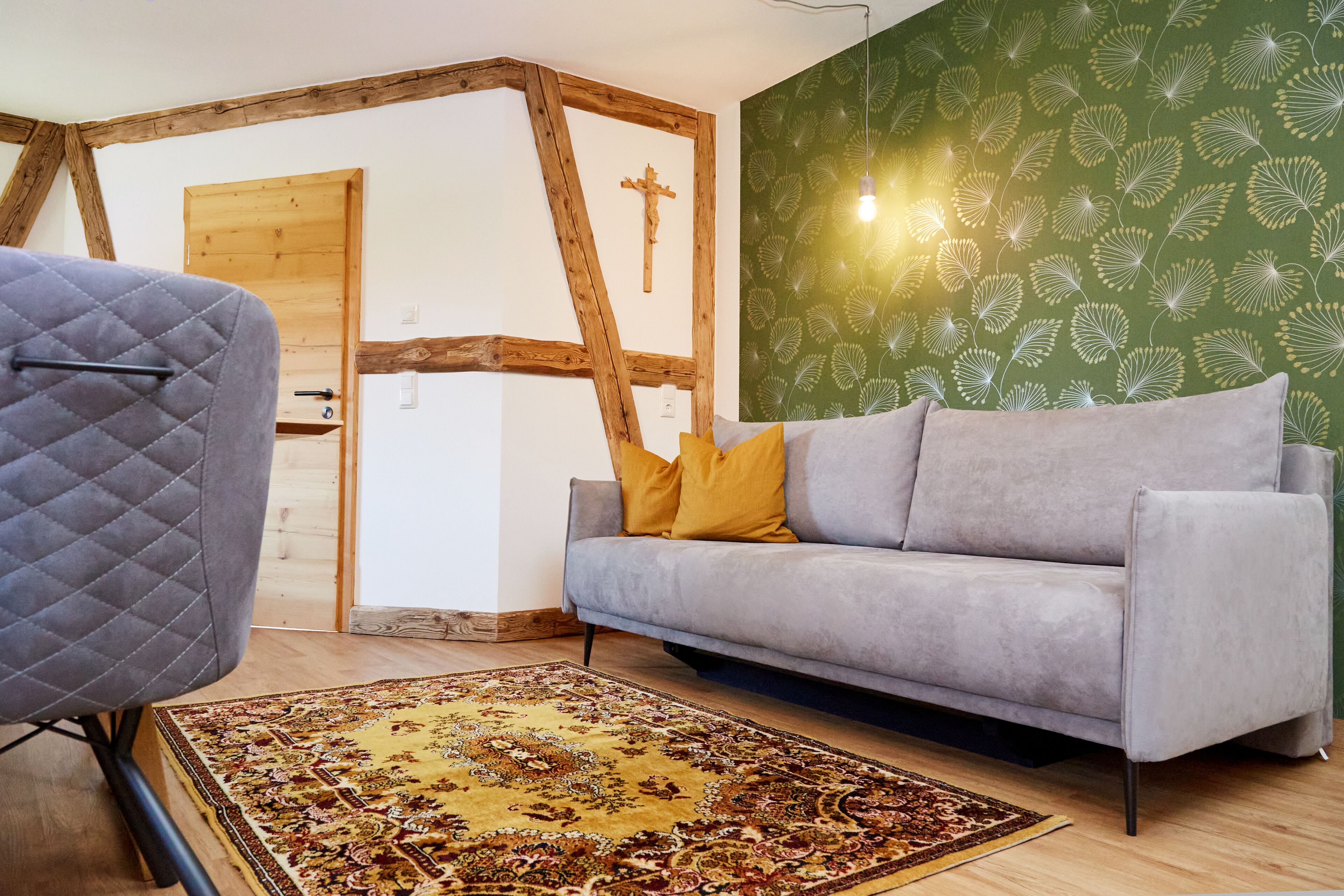 Une pièce avec un canapé gris, un tapis à motifs, des poutres en bois et un mur vert orné d’un motif de feuilles.