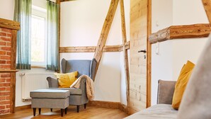 Une pièce chaleureuse avec des poutres apparentes, un fauteuil gris agrémenté d’un coussin jaune, un pouf gris et un lit recouvert d’une couverture grise.