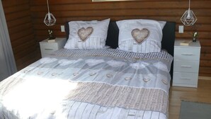 A bedroom with a wooden log wall, a bed with a heart-patterned bedspread, two bedside tables with white lamps, and a painting above the bed.