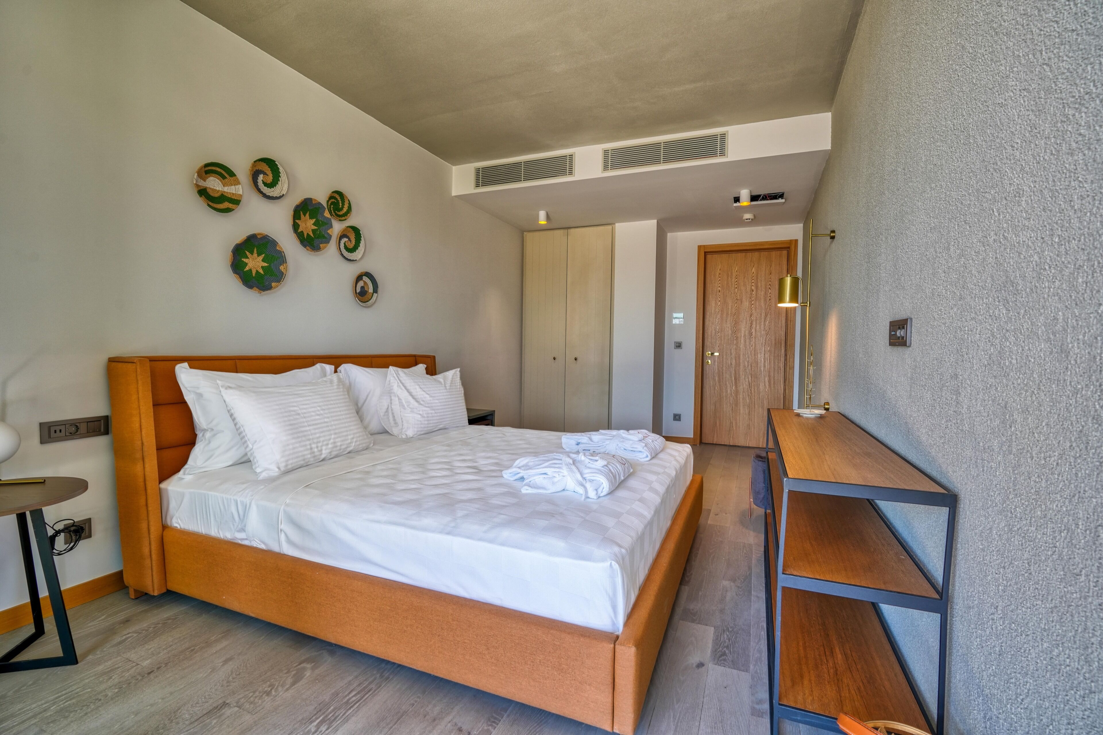 A modern hotel room with a large bed, a wooden headboard, a bedside table, and a small desk with a lamp.