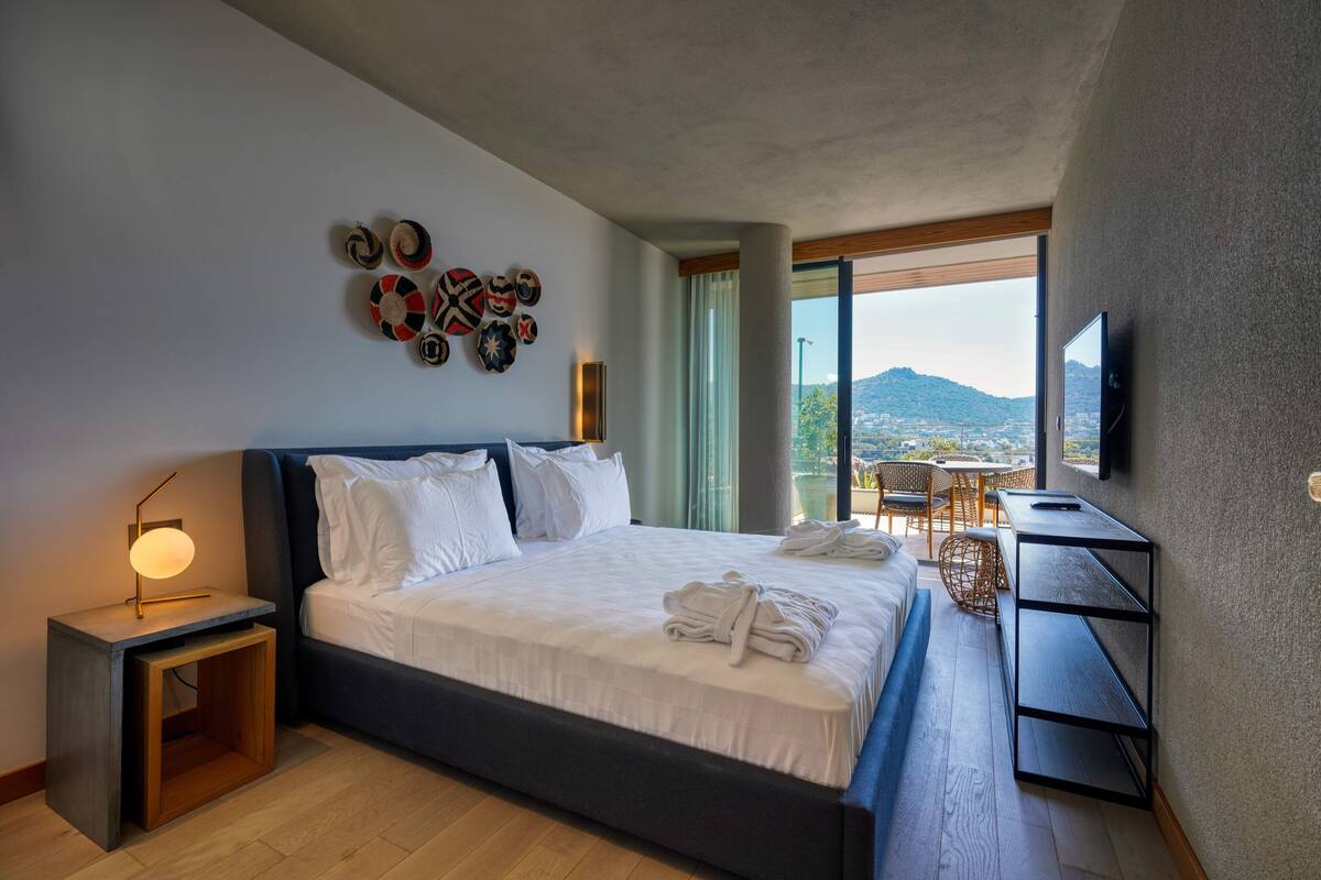 A modern hotel room with a large bed, a bedside table with a lamp, a TV mounted on the wall, and a balcony with a view.