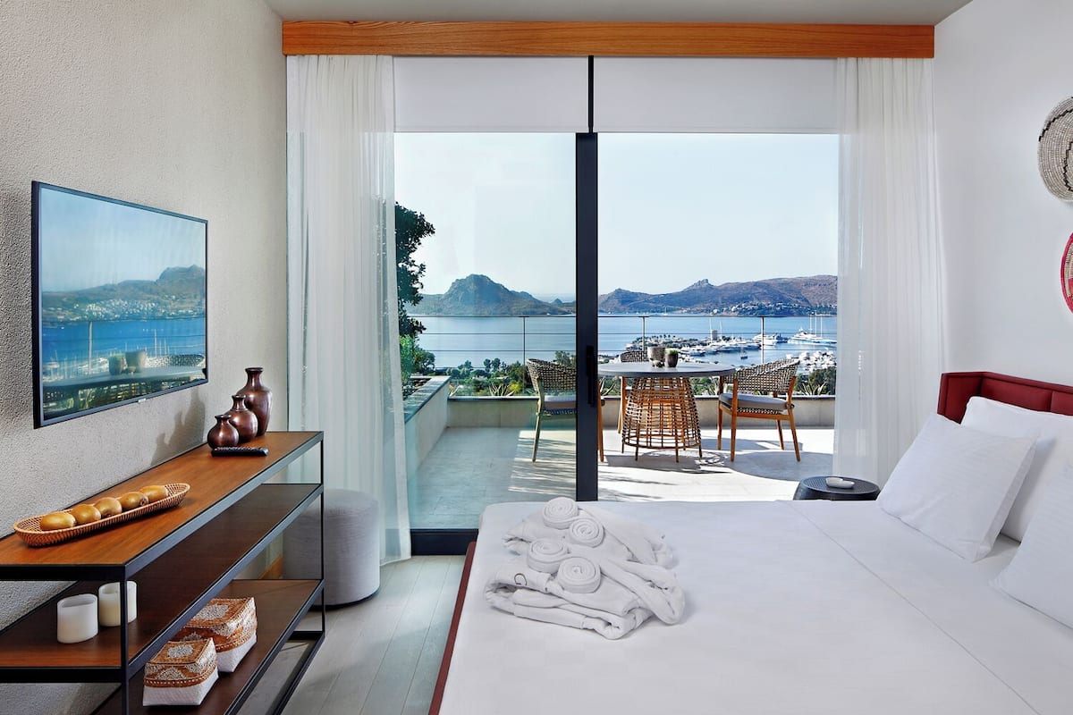 A hotel room with a bed, a desk with a basket of bread, a chair, and a view of the sea and boats.