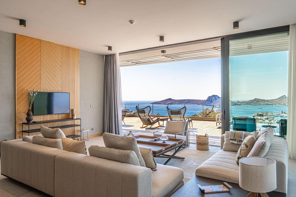 A modern living room with a large flat-screen TV, comfortable seating, and a view of the ocean.
