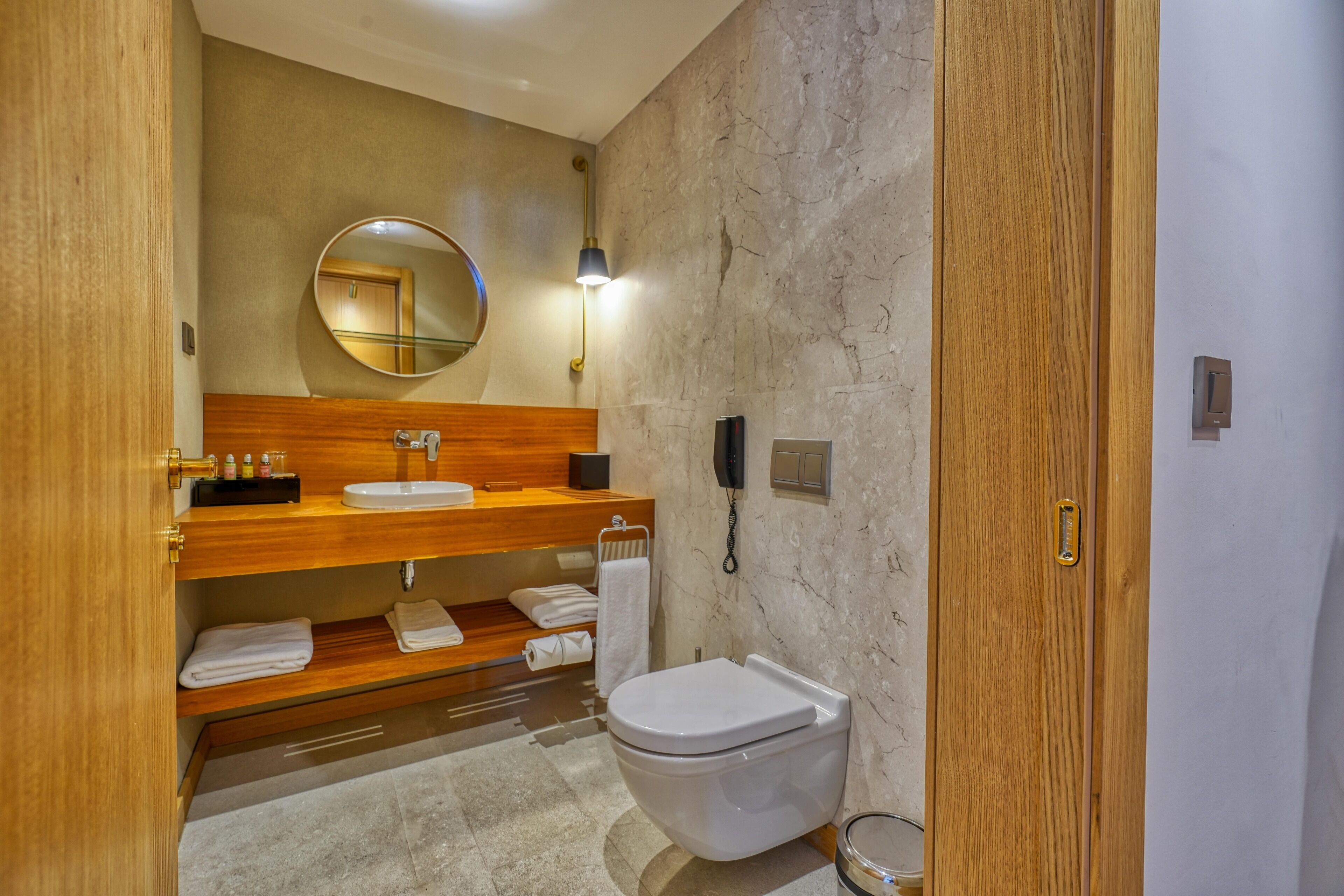 Un baño moderno con tocador de madera, un espejo redondo, un inodoro y una pared de mármol.