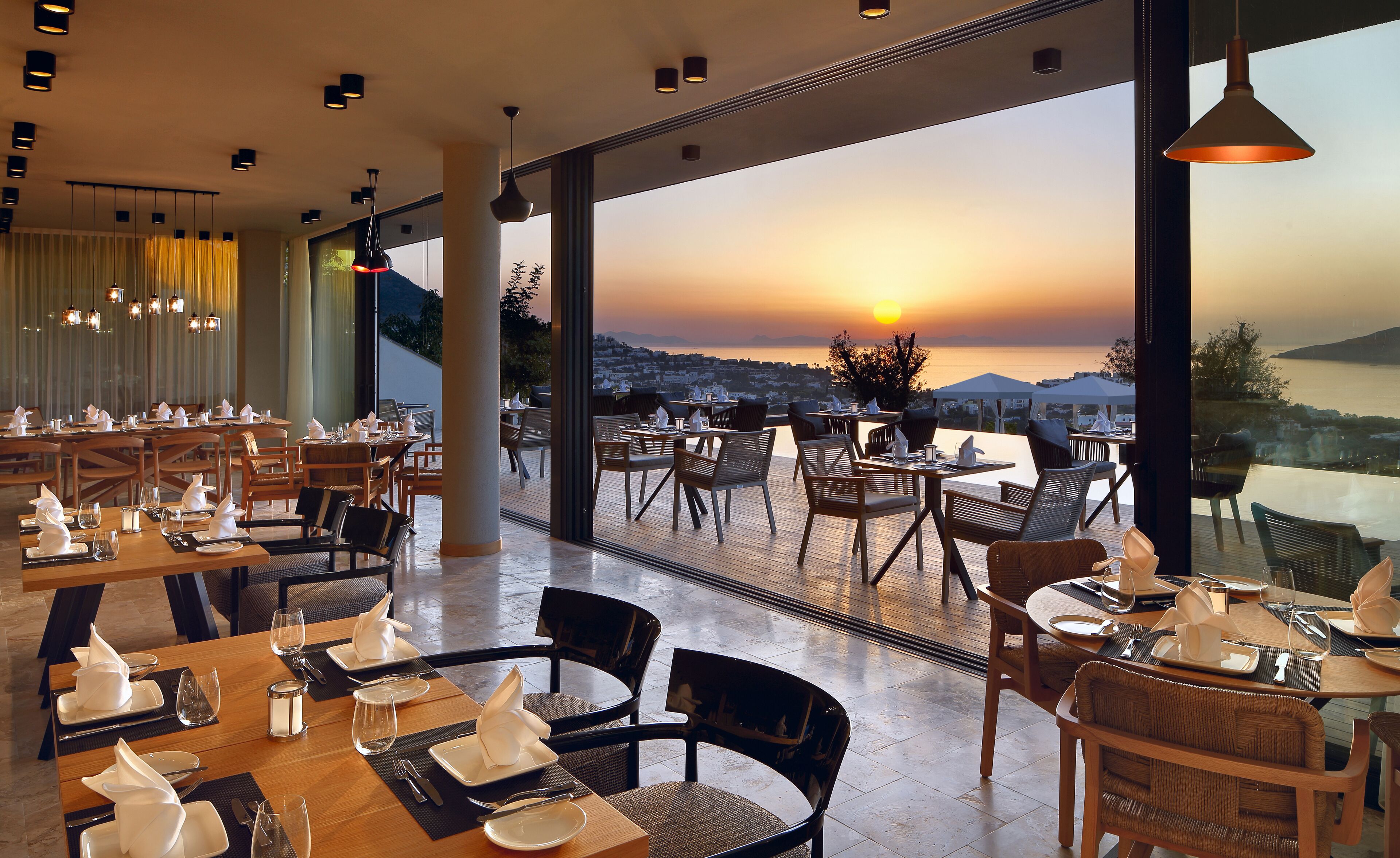 A restaurant with tables set for dining, large windows offering a view of the sunset and landscape.