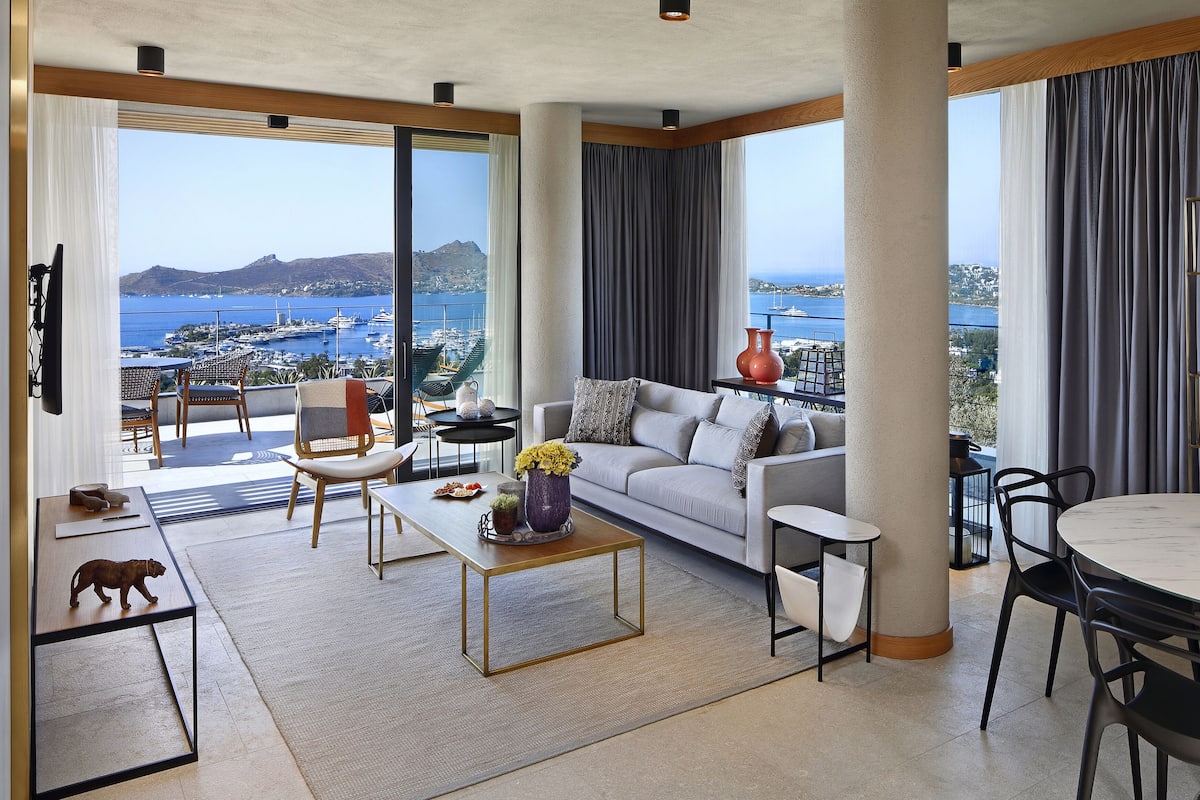 A spacious living room with a large sofa, a coffee table, and a dining area. A balcony with a view of the sea and boats is visible.