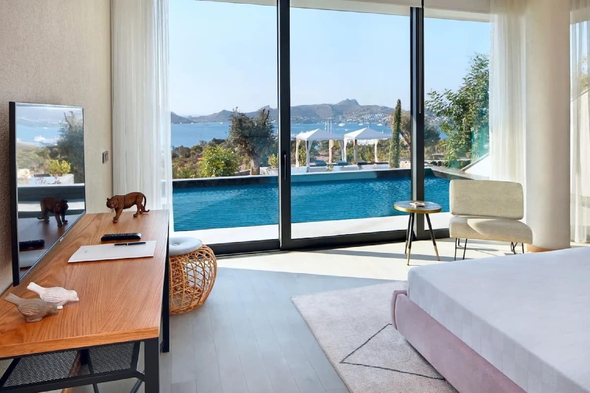 A modern hotel room with a large window, a wooden desk, a bed, and a view of a swimming pool and mountains.