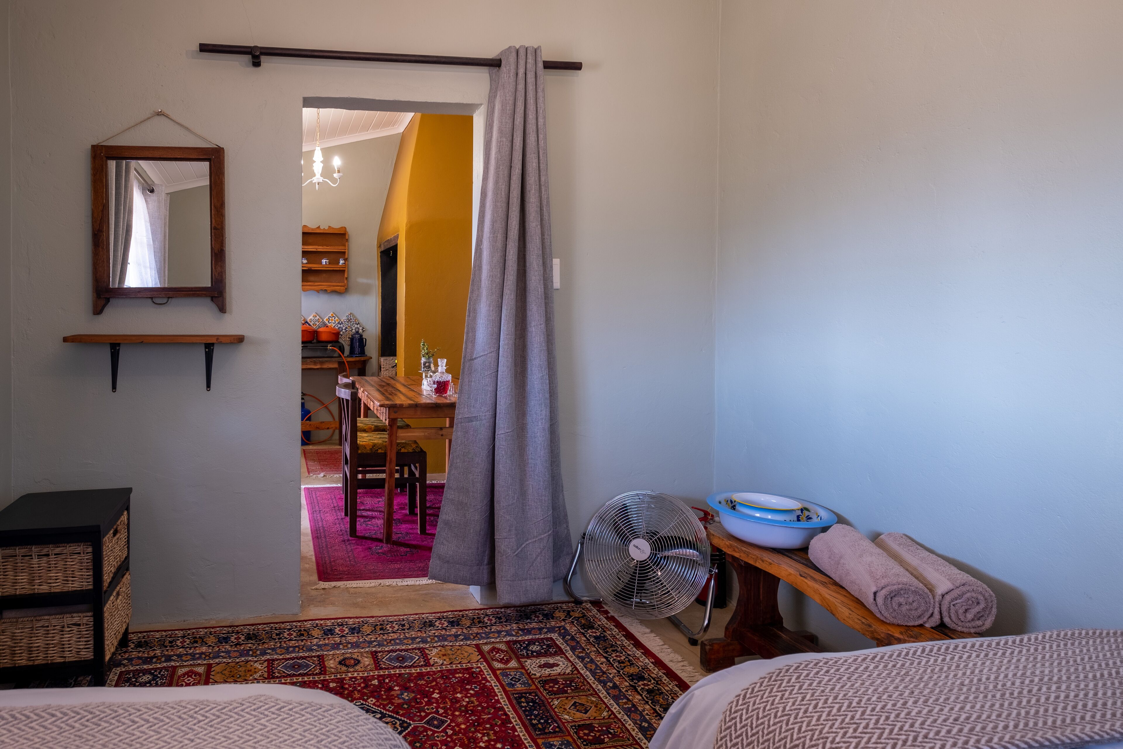 A room with a bed, a fan, a small table with towels, a mirror, and a view into another room with a dining table and chairs.
