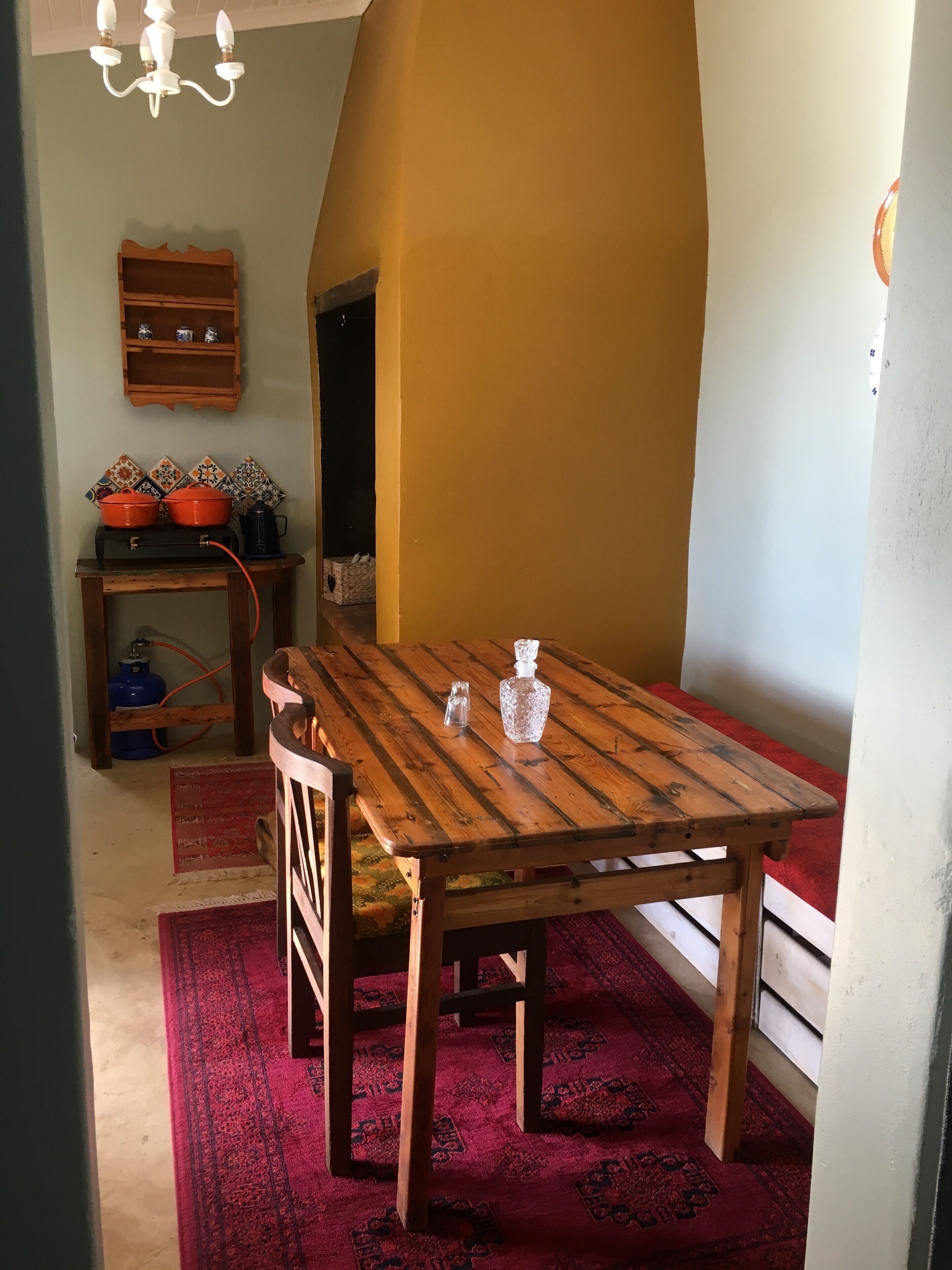 Comedor con mesa de madera, sillas, alfombra roja, lámpara de candelabro y estante con vajilla.